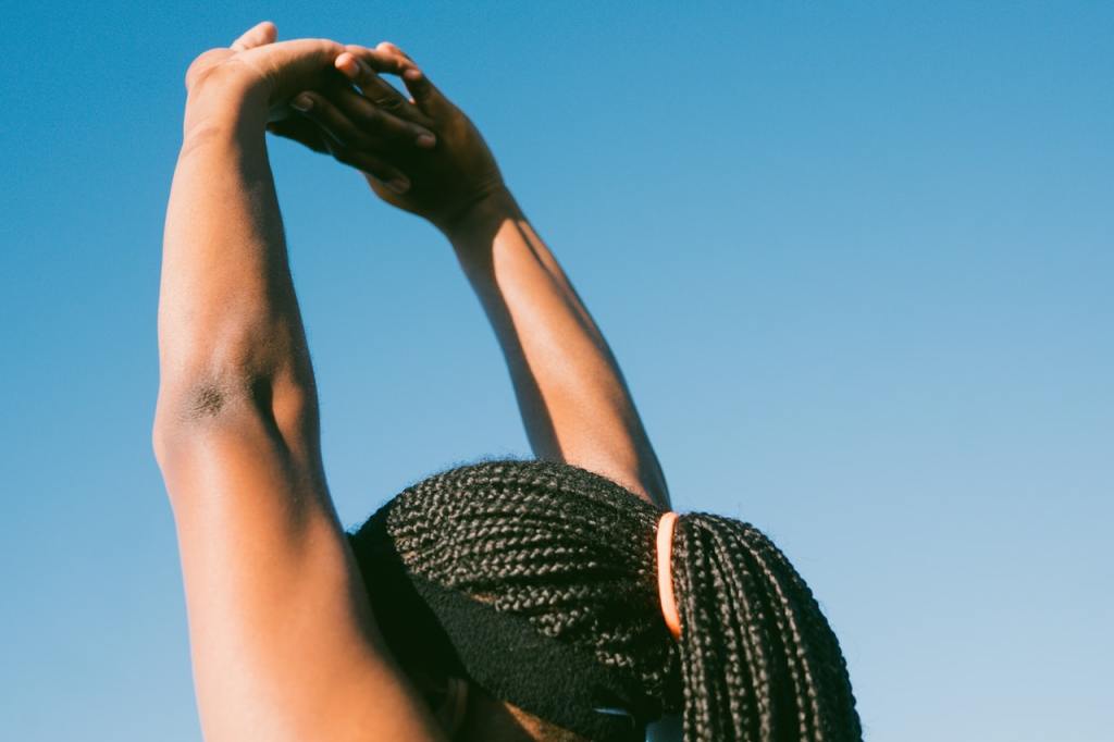 A Black person with long, braided hair stretches into a blue sky.